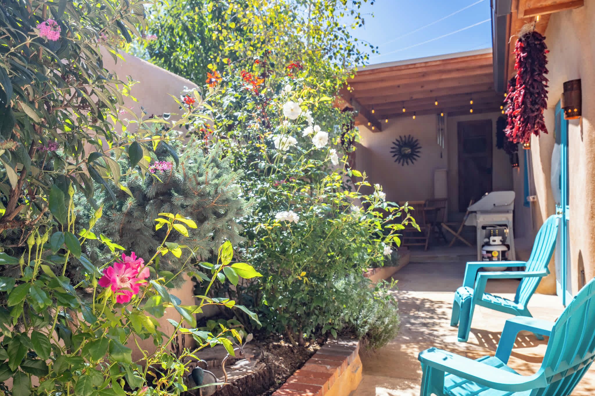 A patio area with a variety of plants and flowers, a barbecue grill, and two blue chairs.