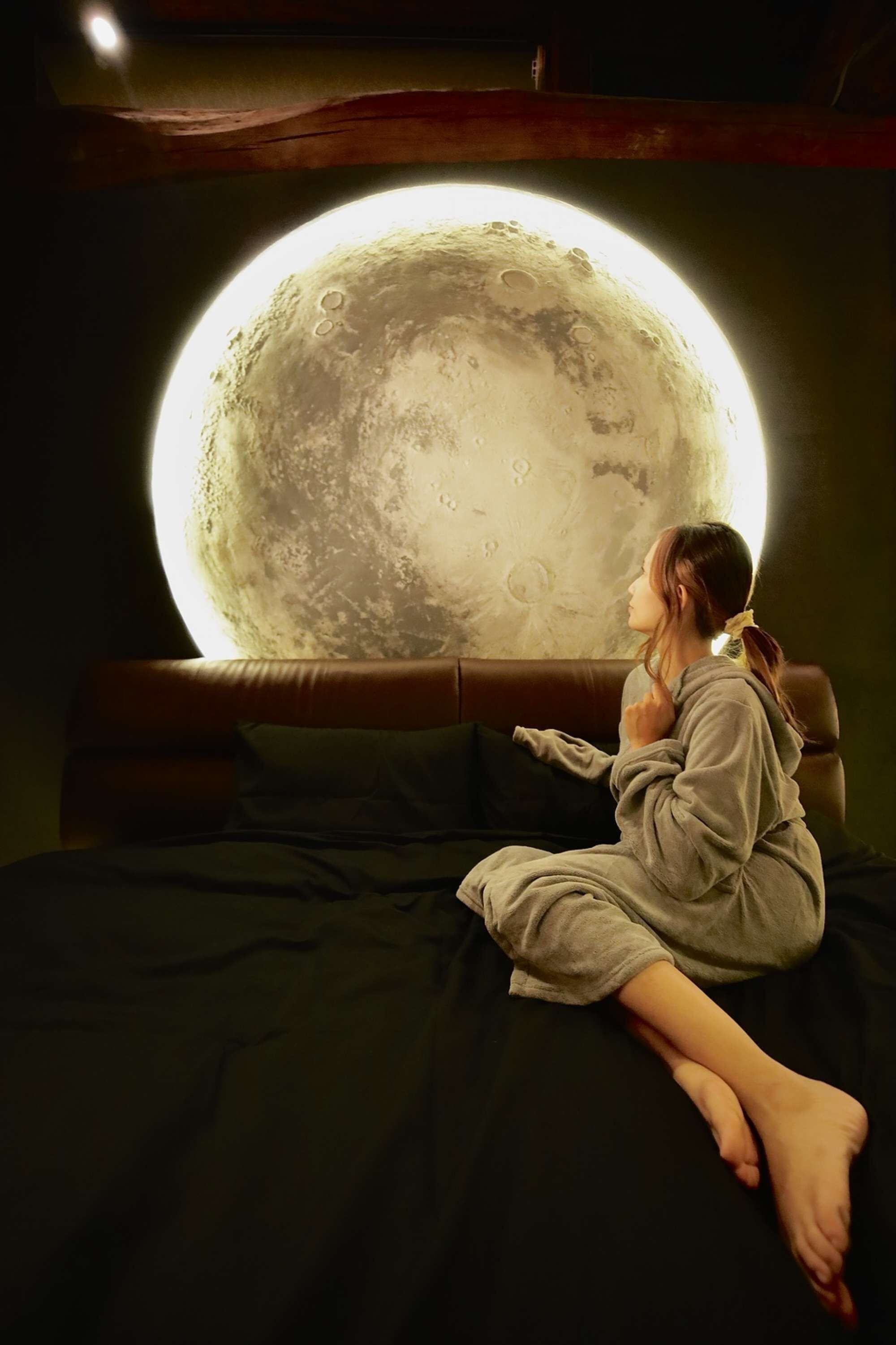 Une personne est assise sur un lit, et en arrière-plan, on aperçoit un grand objet lumineux qui ressemble à une lune.