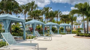 Een zwembadgedeelte van een resort met ligbedden en parasols, omgeven door palmbomen en een heldere lucht.