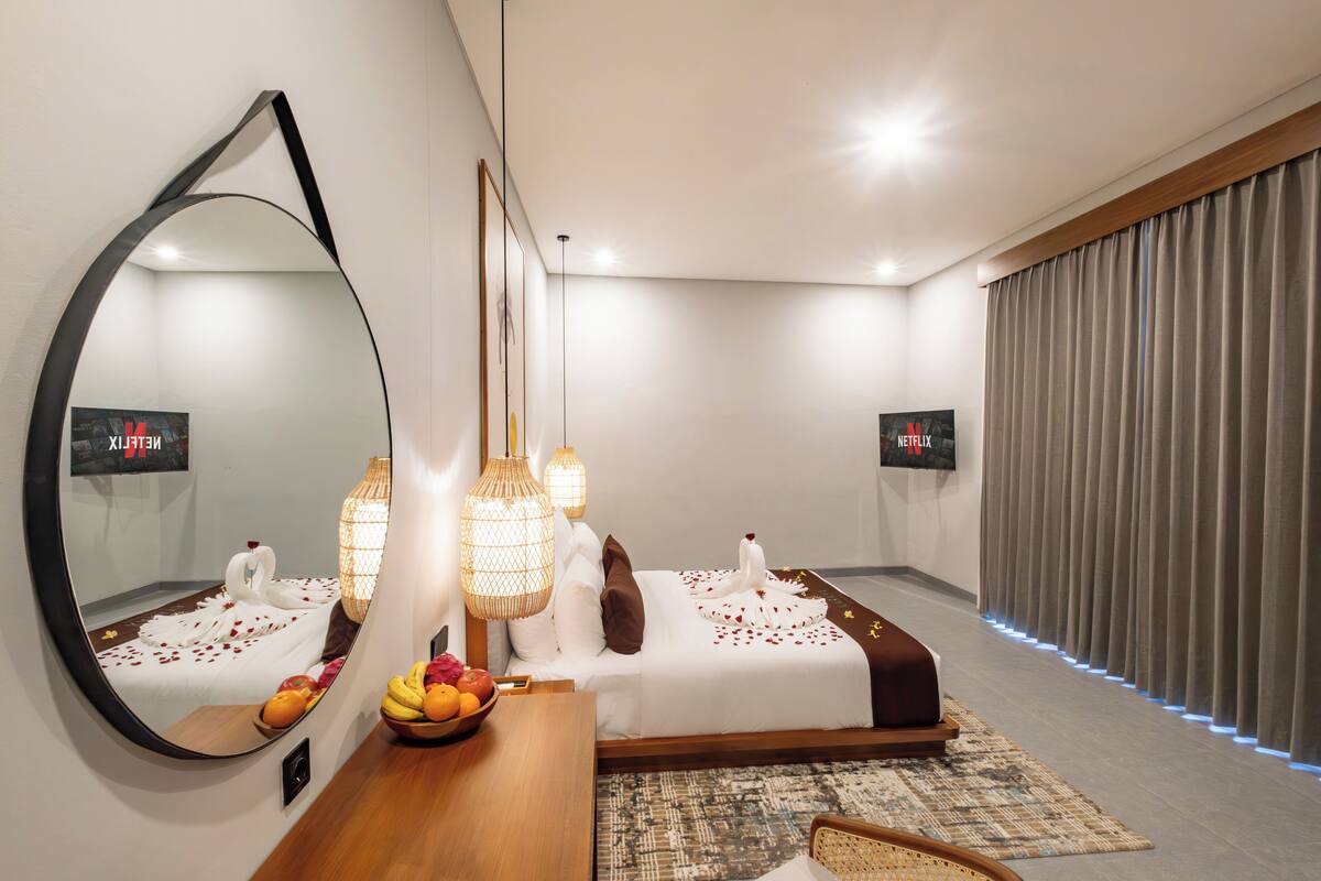 A modern hotel room with a large mirror, a wooden desk with a fruit bowl, a bed with decorative swans and roses, and two Netflix screens.