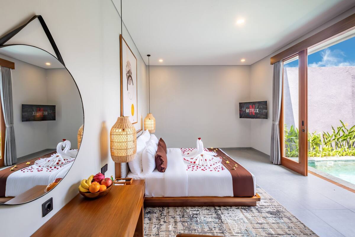A modern hotel room with a large bed, a bedside table with fruit, a mirror, and a view of a swimming pool.