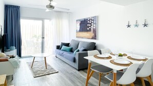 A modern living room with a sofa, dining table, and ceiling fan. - Poolside Paradise by Best Holidays Fuerteventura (Corralejo)