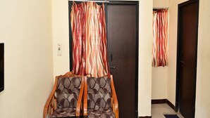 A hotel room with a bed, a wooden chair, a small table, an air conditioner, and a door with striped curtains. - Hotel Dhayalan (Mayiladuthurai)