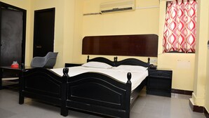 A bedroom with a black bed, a nightstand, a desk, a chair, a wardrobe, and a window with patterned curtains. - Hotel Dhayalan (Mayiladuthurai)