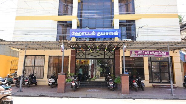 A building with a blue sign, a storefront with a signboard, and several parked motorcycles. - Hotel Dhayalan (Mayiladuthurai)
