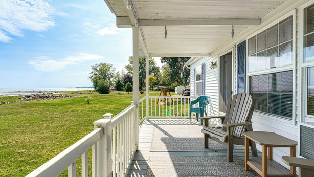 A covered porch with a view of the ocean, a grassy lawn, and a wooden deck with chairs and a table.