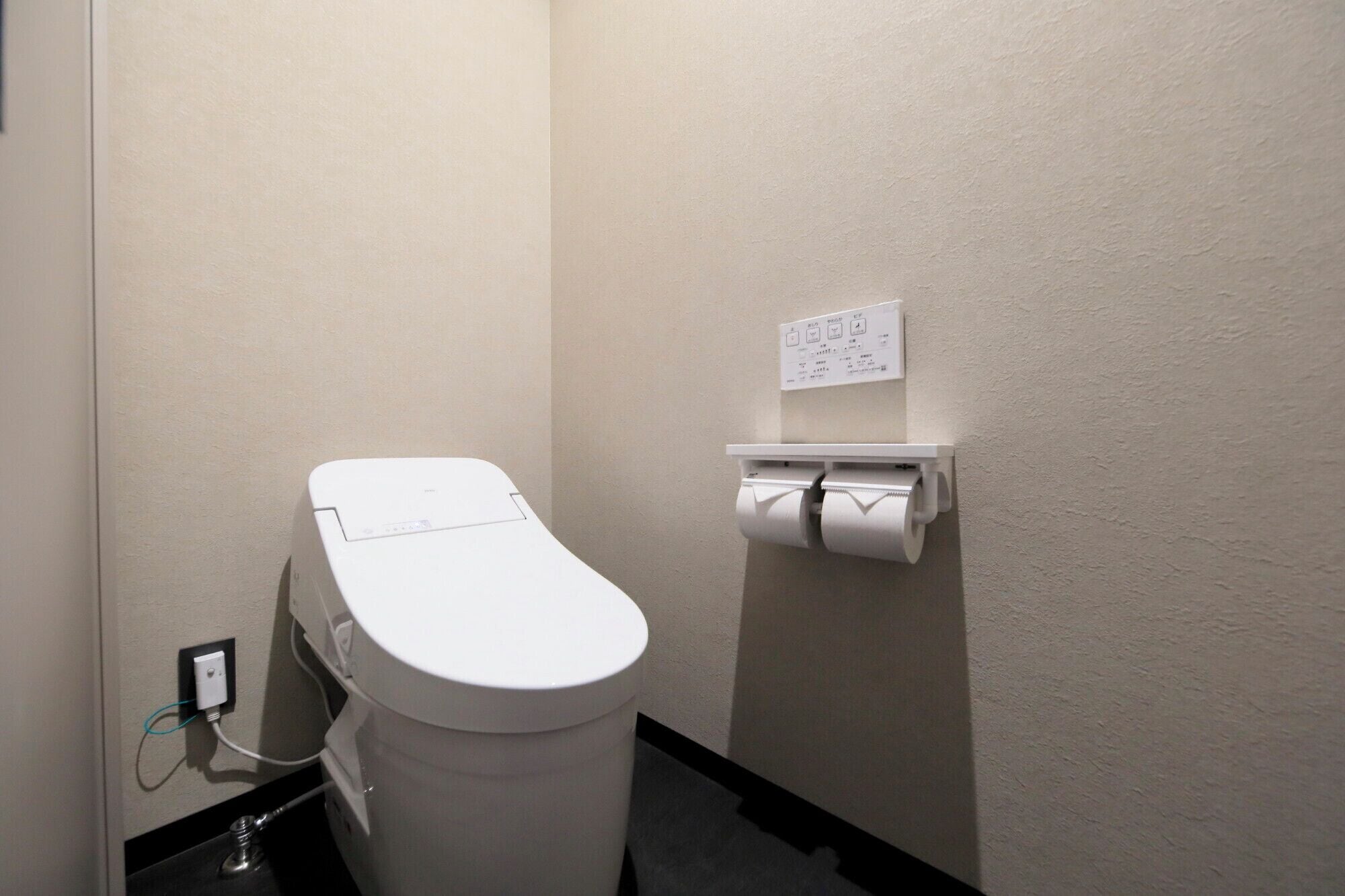 A white toilet with a control panel, a paper towel dispenser, and a power outlet on the wall.