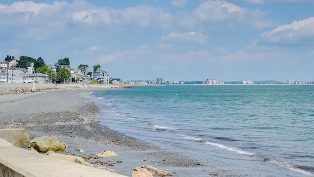 A beach with a concrete walkway, calm water, and buildings in the background. - Steps to Short Beach: Ocean-View Apt w/ Deck! (Winthrop)