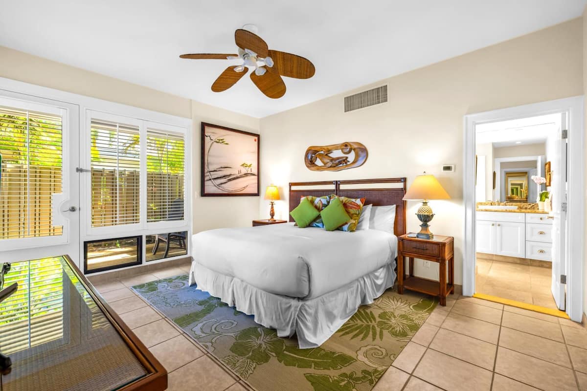 Quarto de hotel com uma cama, duas mesas de cabeceira, um ventilador de teto, uma janela com persianas, um quadro e um banheiro visível através de uma porta aberta.