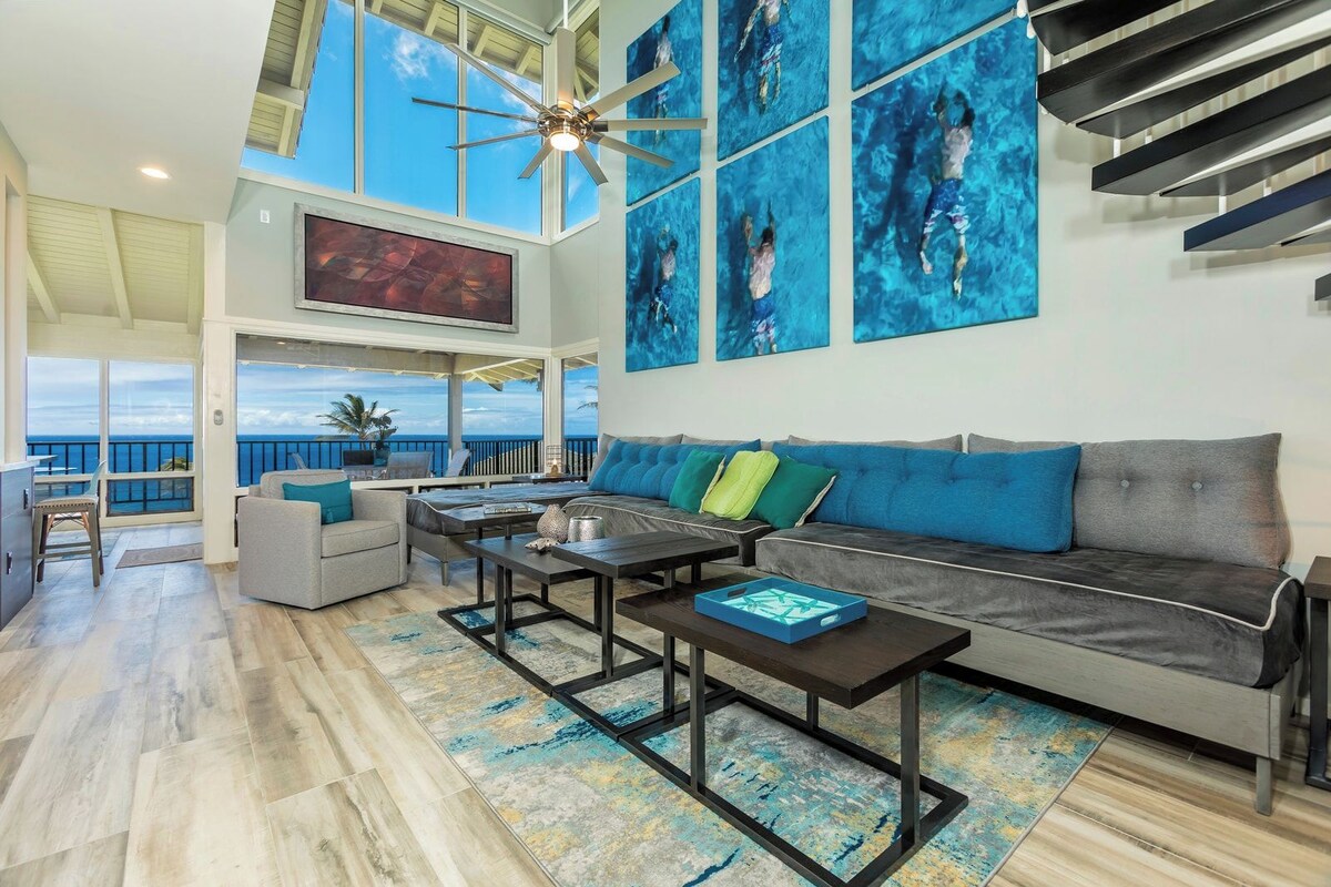 A spacious living area with a large window overlooking the ocean, a ceiling fan, and a modern sofa with colorful cushions.