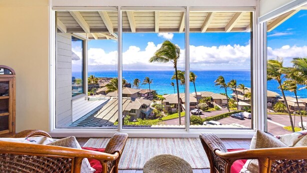 A room with a large glass door leading to a balcony with a view of the ocean and palm trees. - Kapalua Bay Villas Resort by KBM (Kapalua)