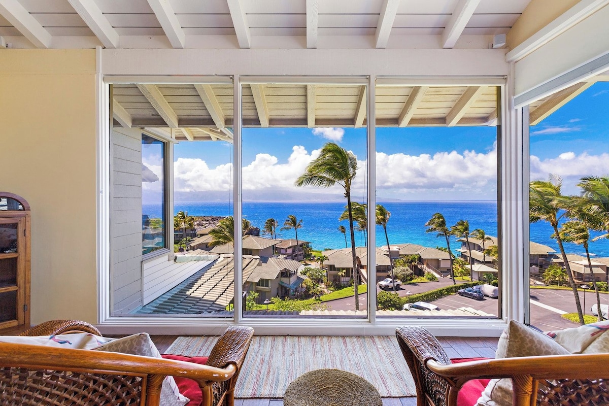 A room with a large sliding glass door leading to a balcony with a view of the ocean and palm trees.