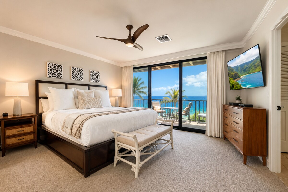 A hotel room with a large bed, a ceiling fan, a TV, a balcony with a view of the ocean, and a desk with a lamp.
