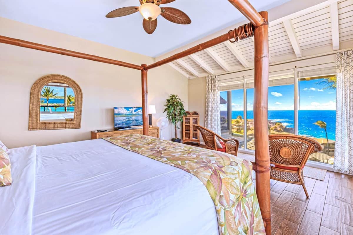 Uma cama de dossel com vista para o oceano, uma televisão sobreposta a um móvel de madeira e cadeiras de vime junto à janela.
