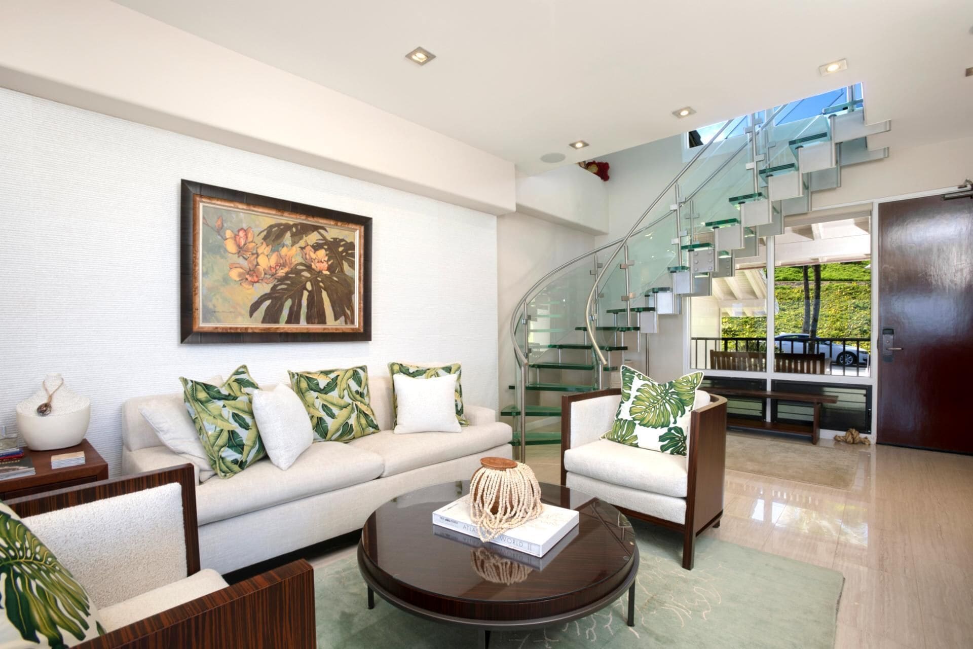 Una sala de estar moderna con un sofá blanco, almohadones estampados verdes, una mesa de centro con un libro y una escalera de cristal.