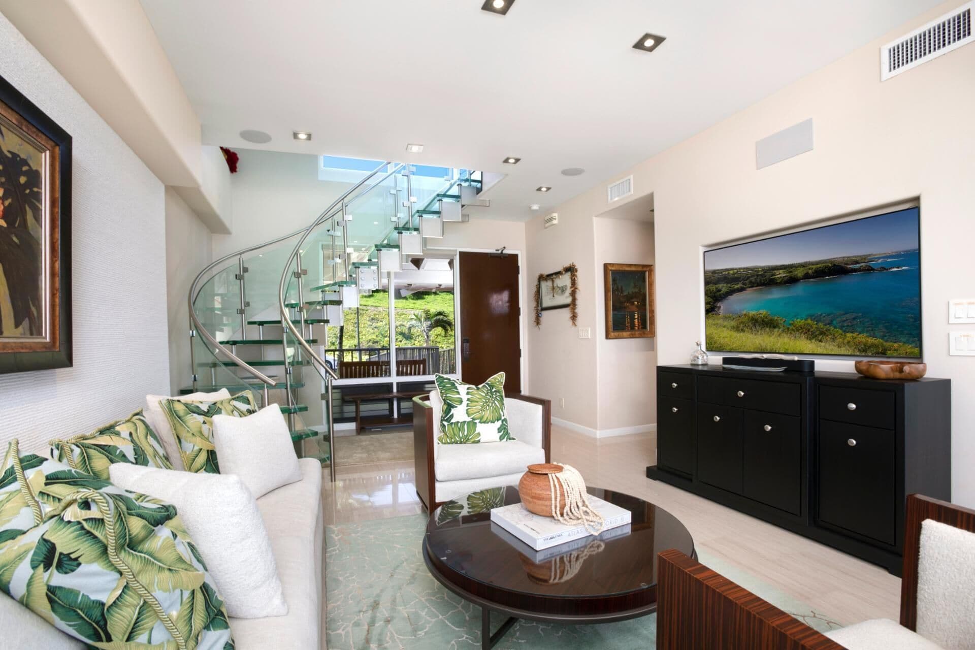 A modern living room with a glass staircase, a flat-screen TV, and a sofa with tropical-themed cushions.