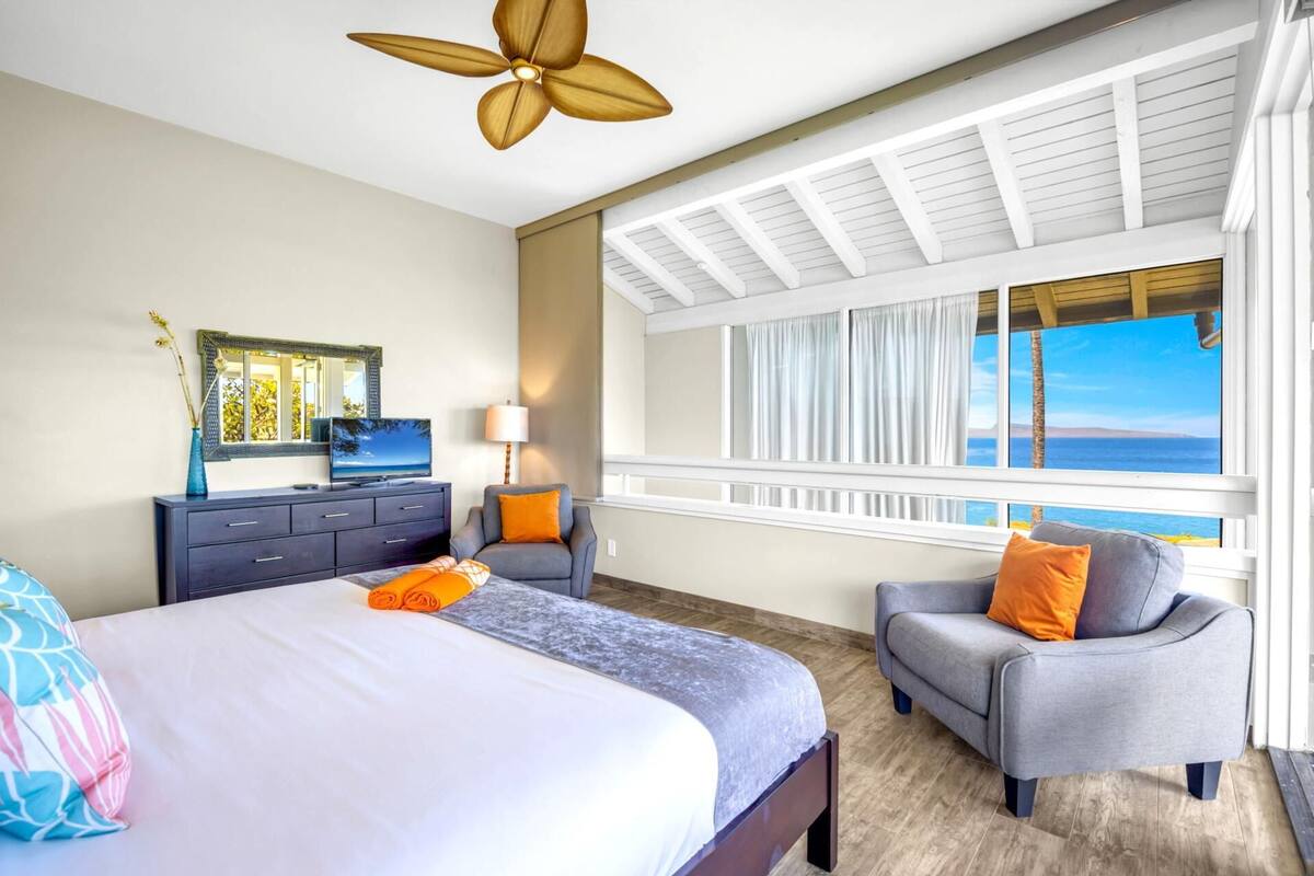 Quarto com cama, escrivaninha, cadeira, janela com vista para o mar e ventilador de teto.