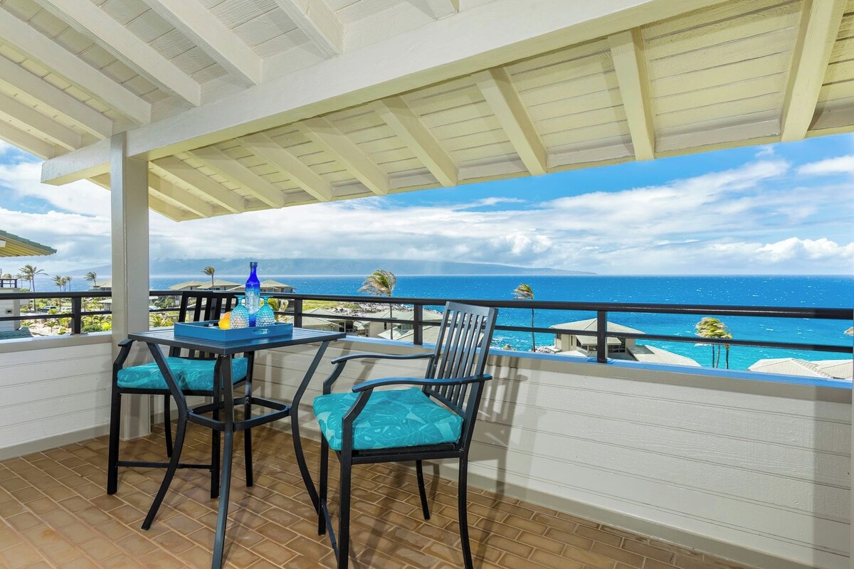 A covered outdoor area with a table and chairs, overlooking a beach with the ocean and palm trees.