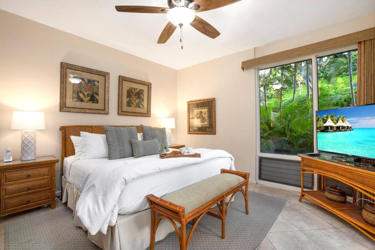Quarto com cama, mesas de cabeceira, televisão e janela com vista tropical.
