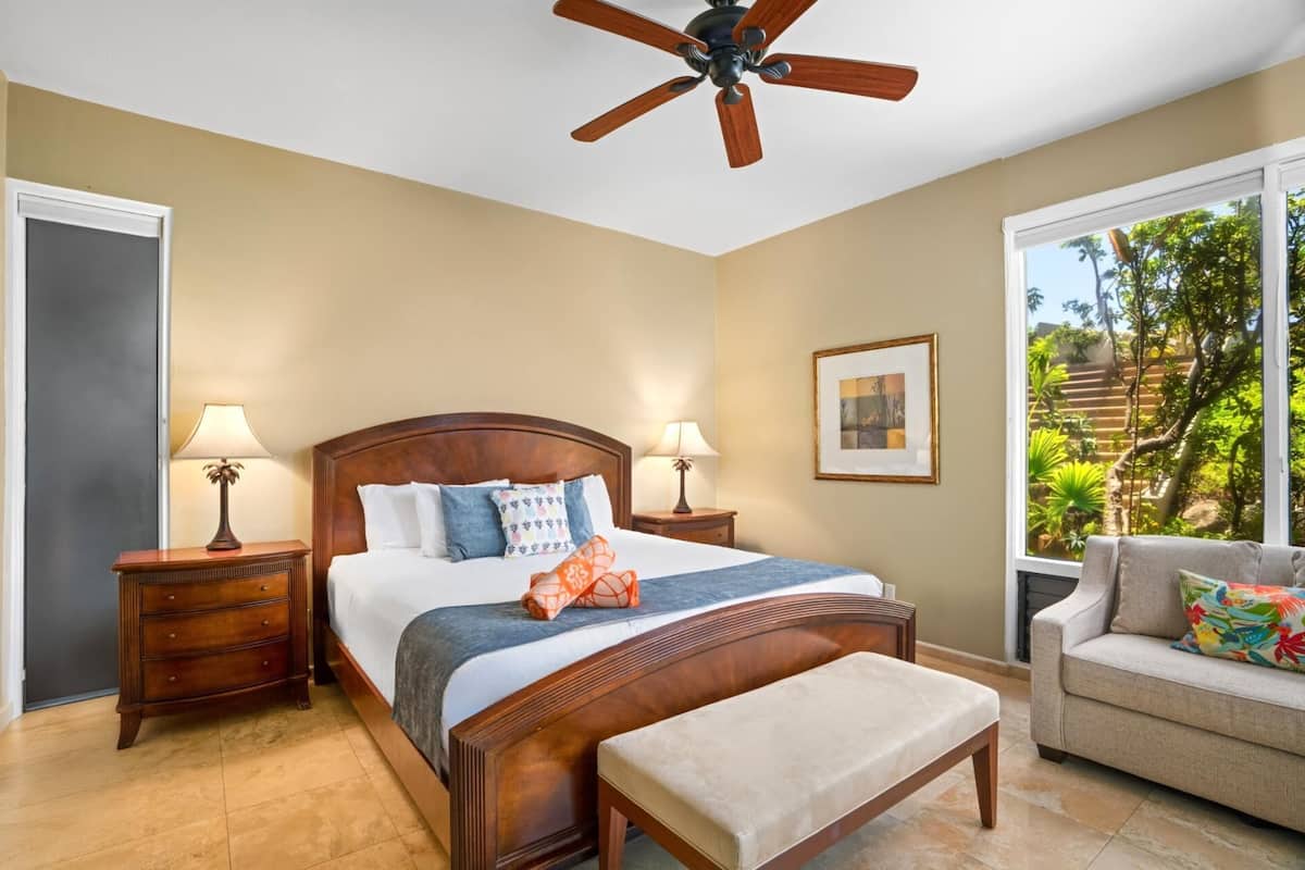 A bedroom with a wooden bed, two bedside tables with lamps, a bench, a window with a view of greenery, and a ceiling fan.