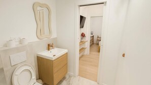 Ein modernes Badezimmer mit Toilette, Waschbecken und Spiegel. Darunter befindet sich ein Holzschrank und zur Rechten ein Duschbereich.