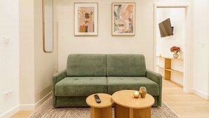 Ein grünes Sofa, zwei gerahmte Kunstwerke, ein Fernseher und ein kleiner Tisch mit Fernbedienung und Kerze.