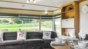 A modern interior with a sofa facing a large window, a dining table set with plates and glasses, and a wooden shelf with decorative items.