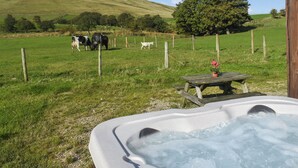 Uma banheira de hidromassagem com uma mesa de madeira e um vaso de flores, cercada por um campo gramado com vacas e uma árvore ao fundo.