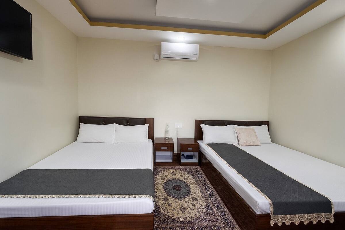A hotel room with two single beds, a small bedside table with a lamp, a wall-mounted air conditioner, and a patterned rug.