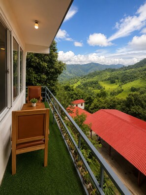 A balcony with a wooden chair and table, potted plants, and a view of green hills and a red-roofed building.