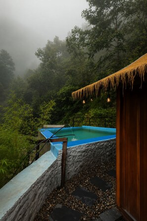 Ein Pool mit einer Holzterrasse, umgeben von üppiger Vegetation und einer Reetdachkonstruktion.