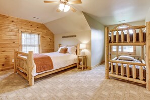 A wooden cabin bedroom with a bunk bed, a single bed, a ceiling fan, and a small bedside table with a lamp.