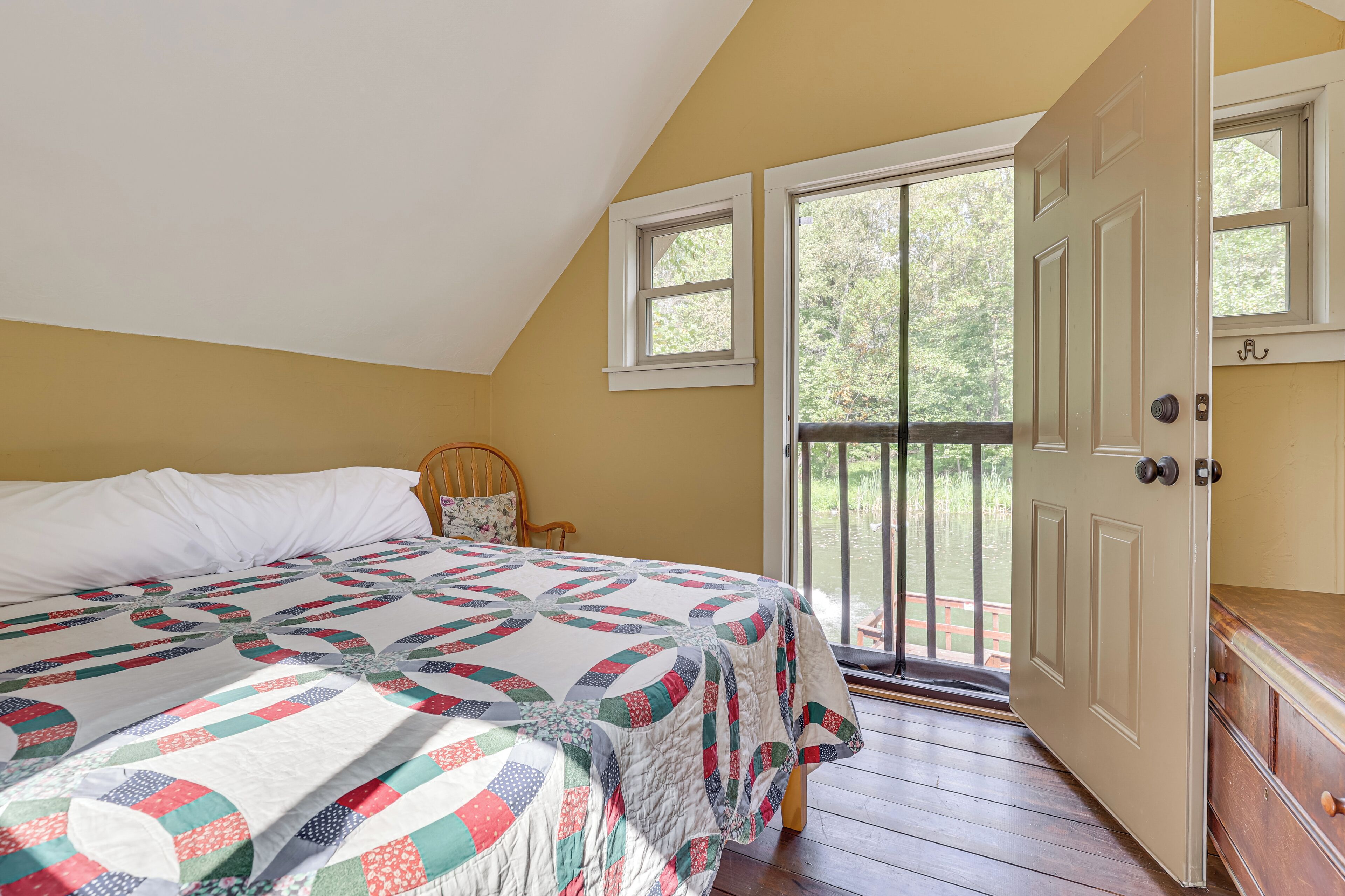 Ein Schlafzimmer mit einem großen Bett, einem Holzstuhl, einer Tür, die auf einen Balkon führt, und Blick auf Bäume.