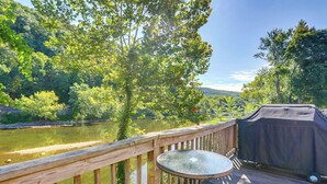 A wooden deck with a glass table overlooking a river, surrounded by lush greenery and a covered barbecue area. - Franklin Home w/ Deck, Creek Views: Walk to Shops (Franklin)