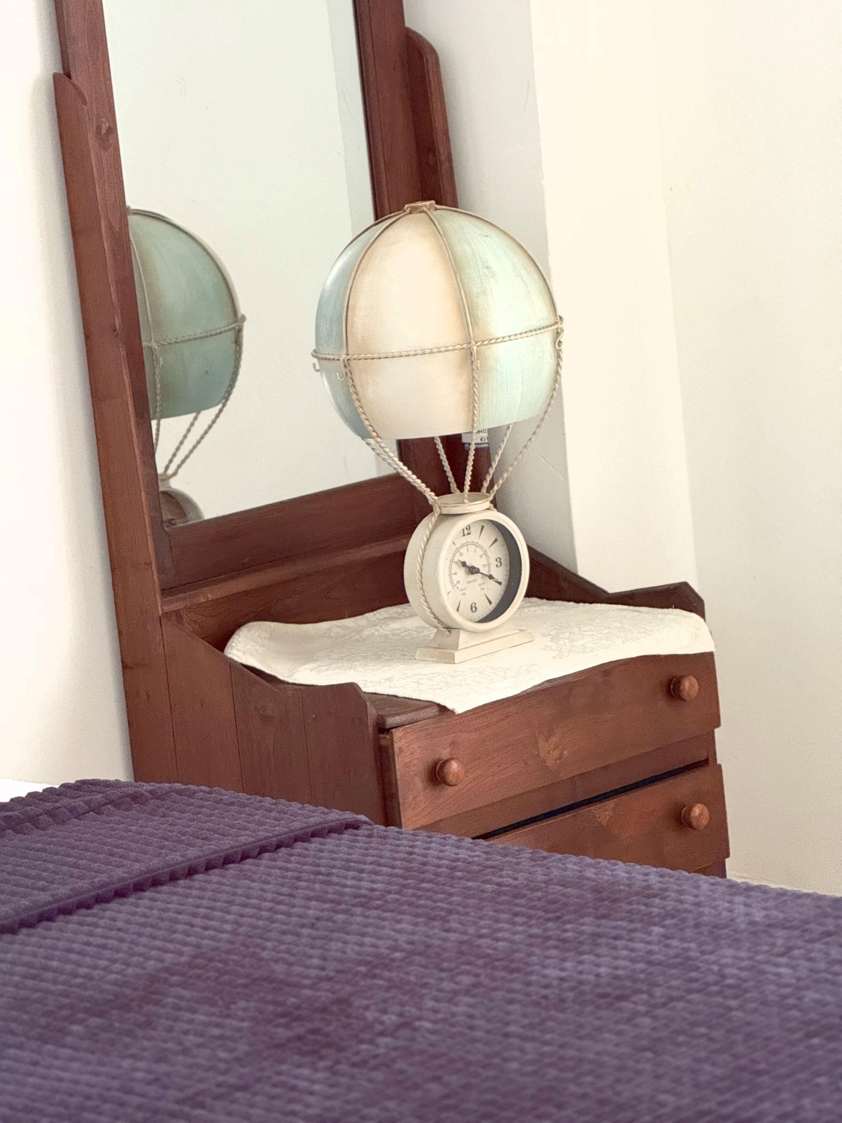 A wooden dresser with a globe and clock on top, a mirror above, and a purple blanket on the bed.