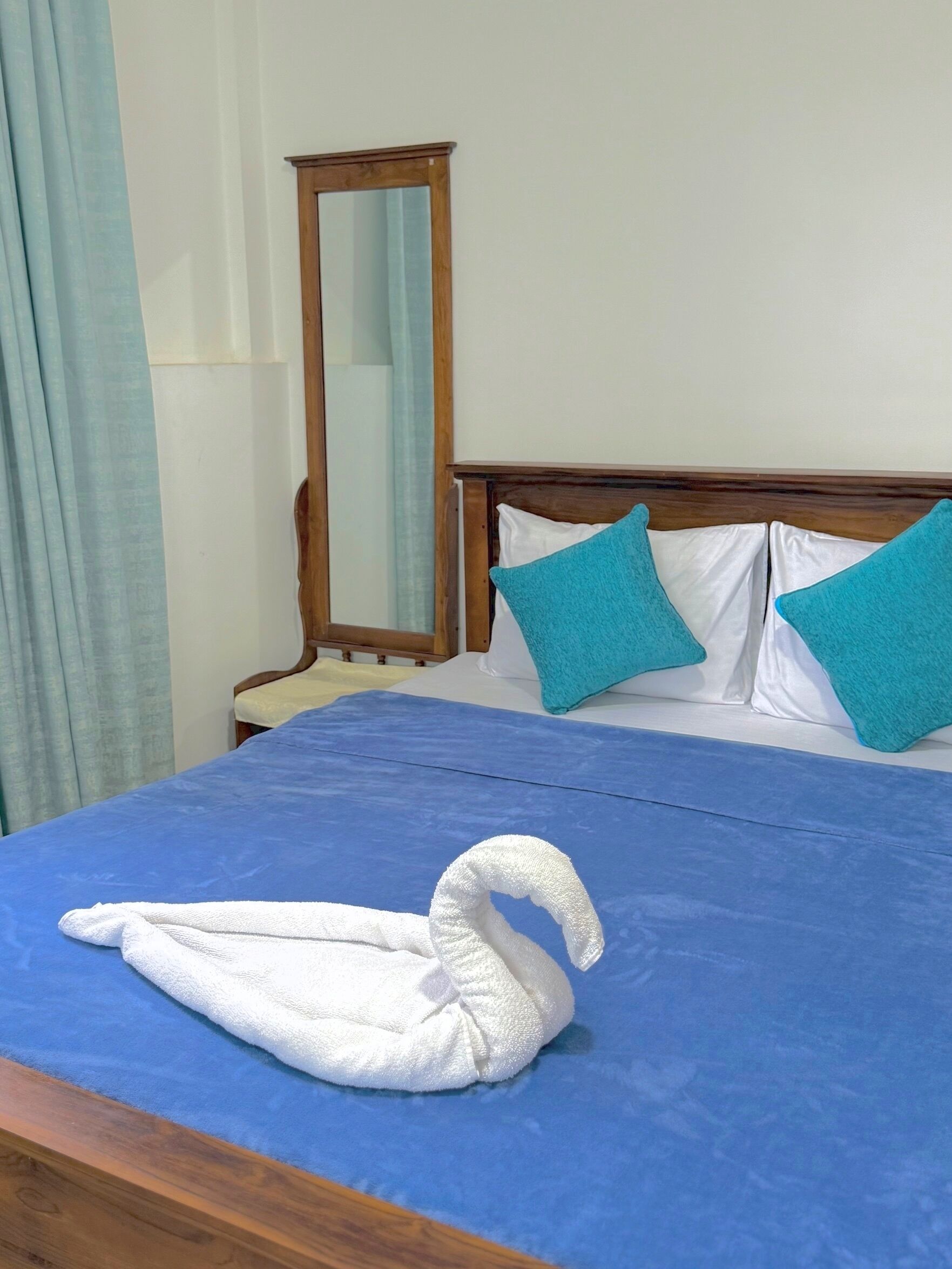 A neatly made bed with a blue bedspread, two white pillows, and two teal pillows. A white towel is folded on the bed.