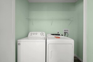 A laundry room with two white washing machines, a shelf above, and a power outlet on the wall. - Lakefront Luxury Retreat | Dock, Kayak & Views (Crossville)