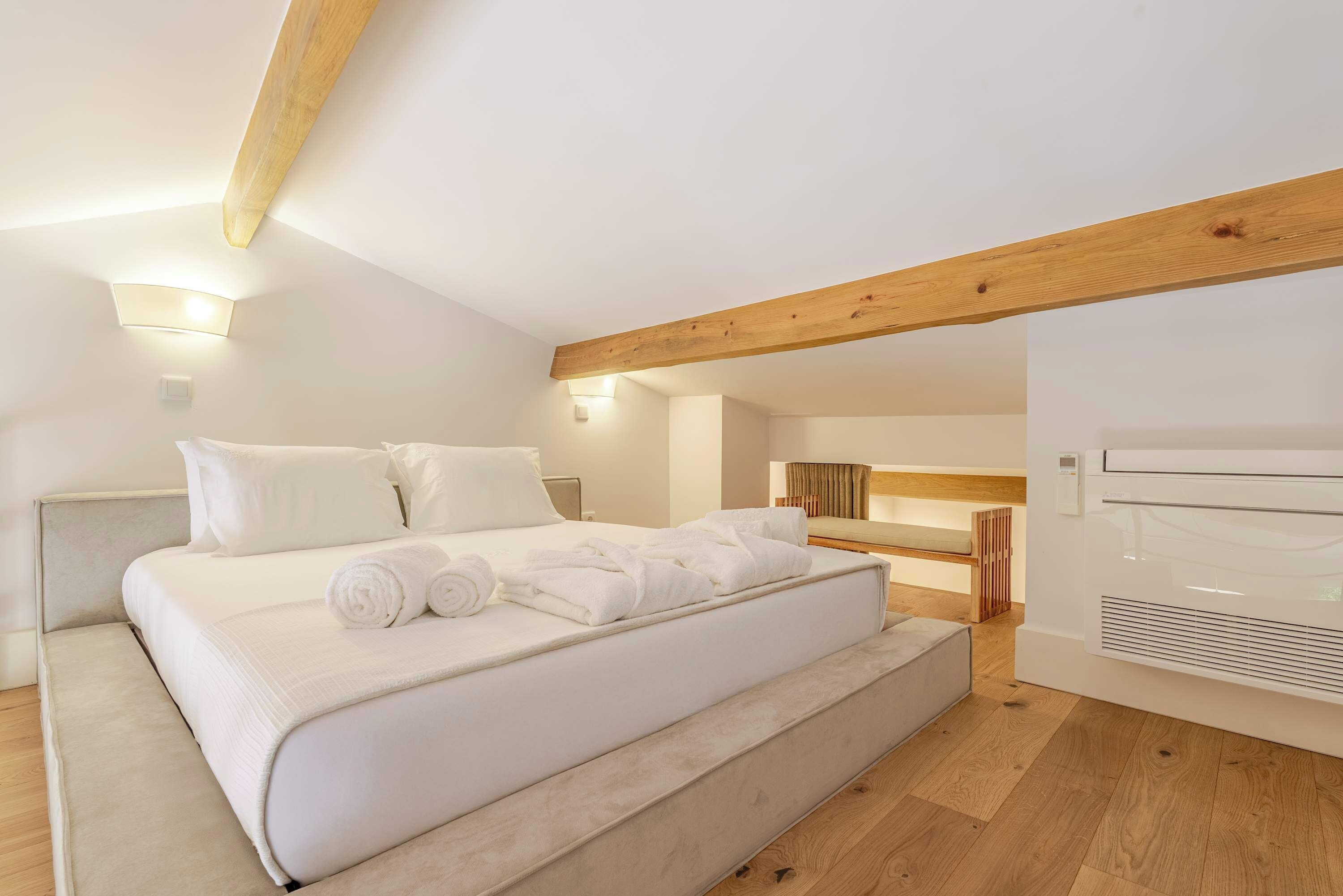 A modern bedroom with a large bed, wooden beams, and a built-in heater.