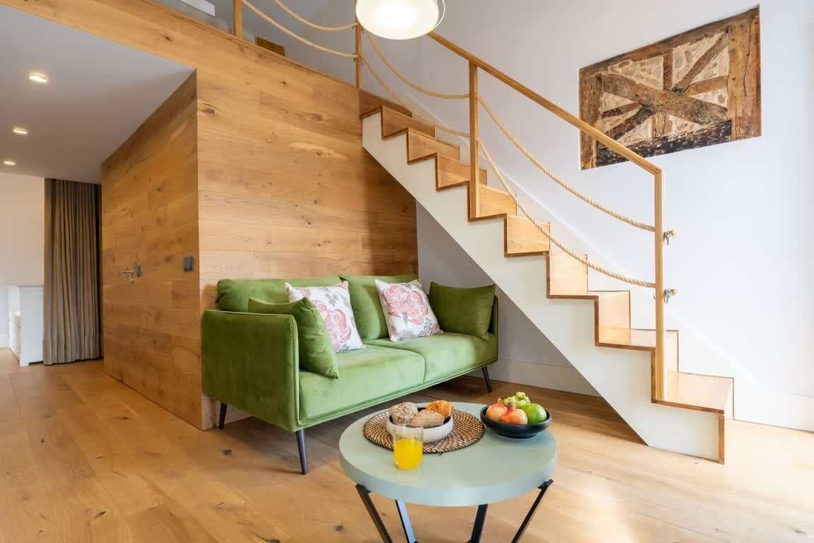 A modern living room with a wooden staircase, a green sofa, and a round table with fruits and a drink.