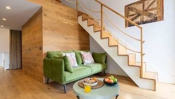 A modern living room with a wooden staircase, a green sofa, and a round table with fruits and a drink.