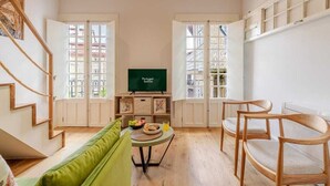 Una sala de estar moderna con una escalera, un sofá verde, sillas de madera, una mesa de comedor con frutas y un vaso, y un televisor sobre un mueble.