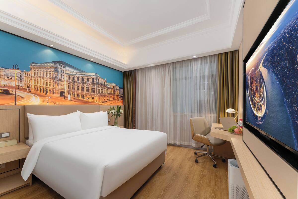 A hotel room with a large bed, a desk with a laptop, a TV, and a mural of a cityscape.