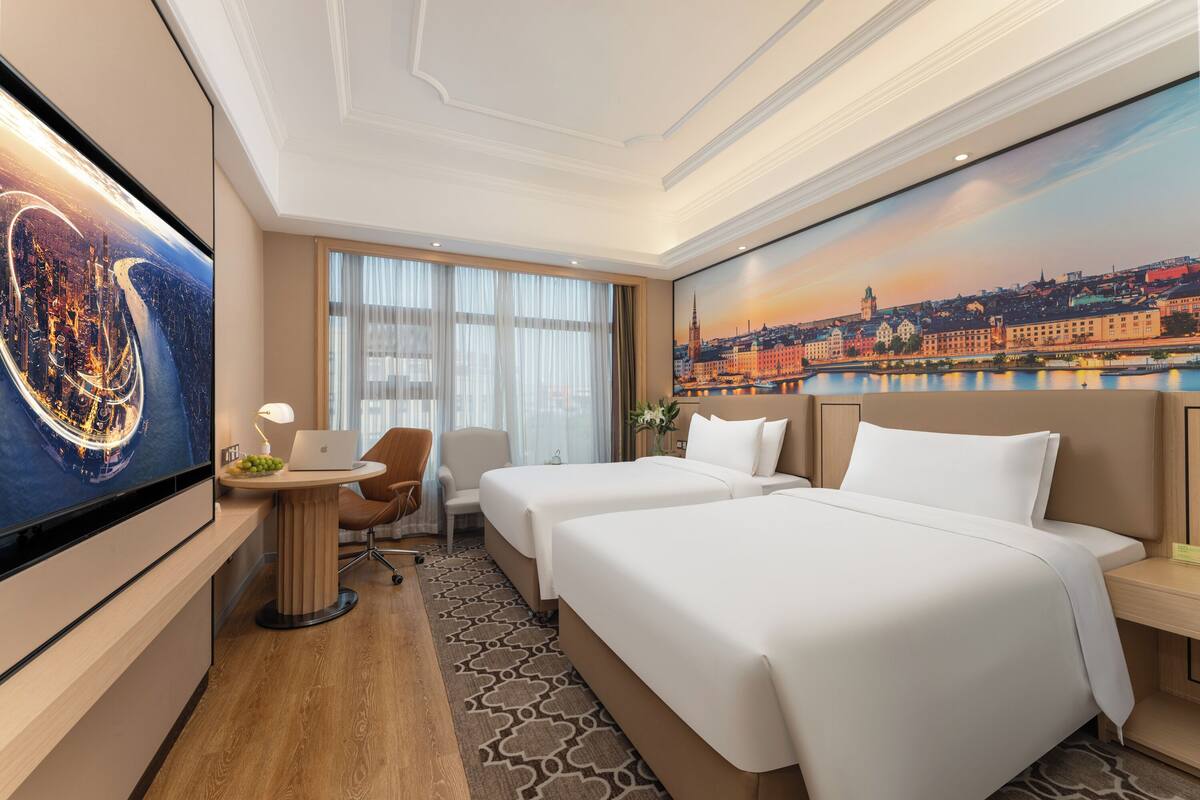 A hotel room with two beds, a desk, a chair, a TV, and a large window with curtains.