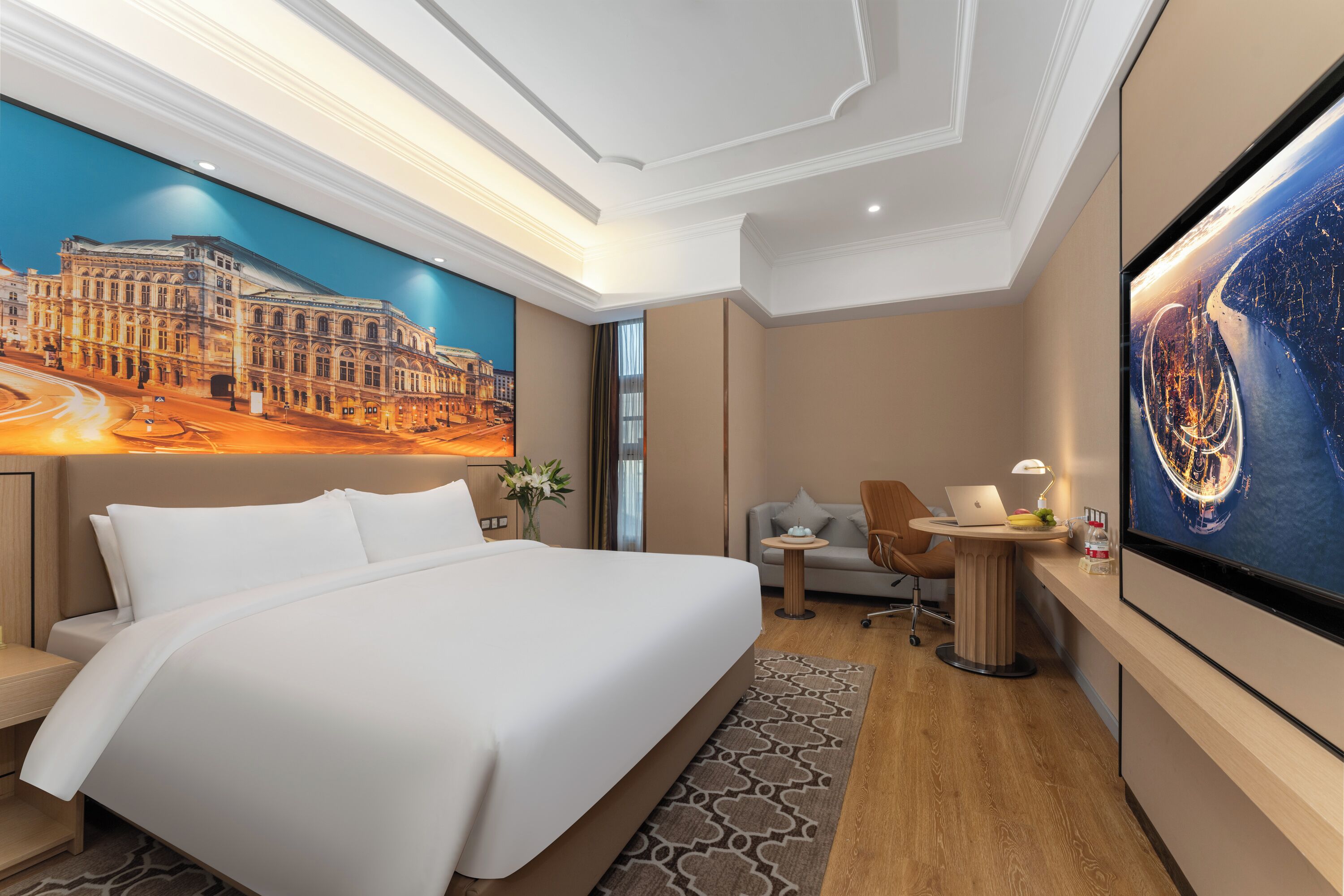 A hotel room with a large bed, a desk with a laptop, a TV, and a mural of a building.