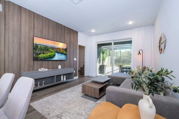 Ein modernes Wohnzimmer mit Flachbildfernseher, grauen Sofas, einem Couchtisch und einem großen Fenster mit Vorhängen.