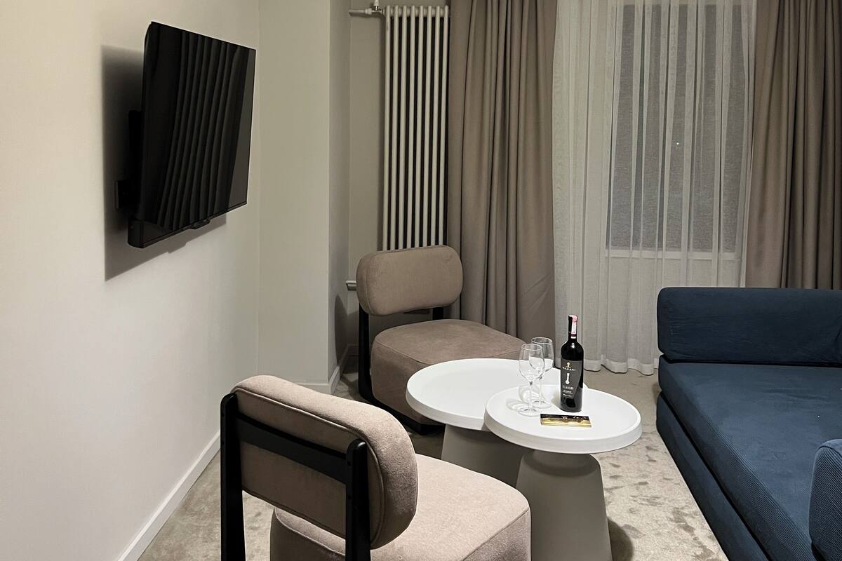 Uma sala de estar moderna com sofá, poltrona e mesa de centro. Uma televisão de tela plana está instalada na parede e há um radiador visível.