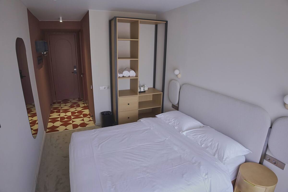 Quarto de hotel com cama, guarda-roupa, espelho, mesa pequena e lixeira.
