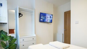 Hotellihuone, jossa on sänky, lipasto, peili, ruukkukasvi, televisio, jolta näkyy Disney-kanava, ja puinen ovi.