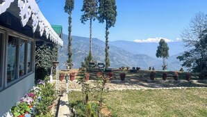 A building with a distinctive white facade and a green roof, surrounded by a garden with potted plants and a clear view of snow-capped mountains in the background. - YuleTea Retreat A Premium Heritage Hotel (Darjeeling)