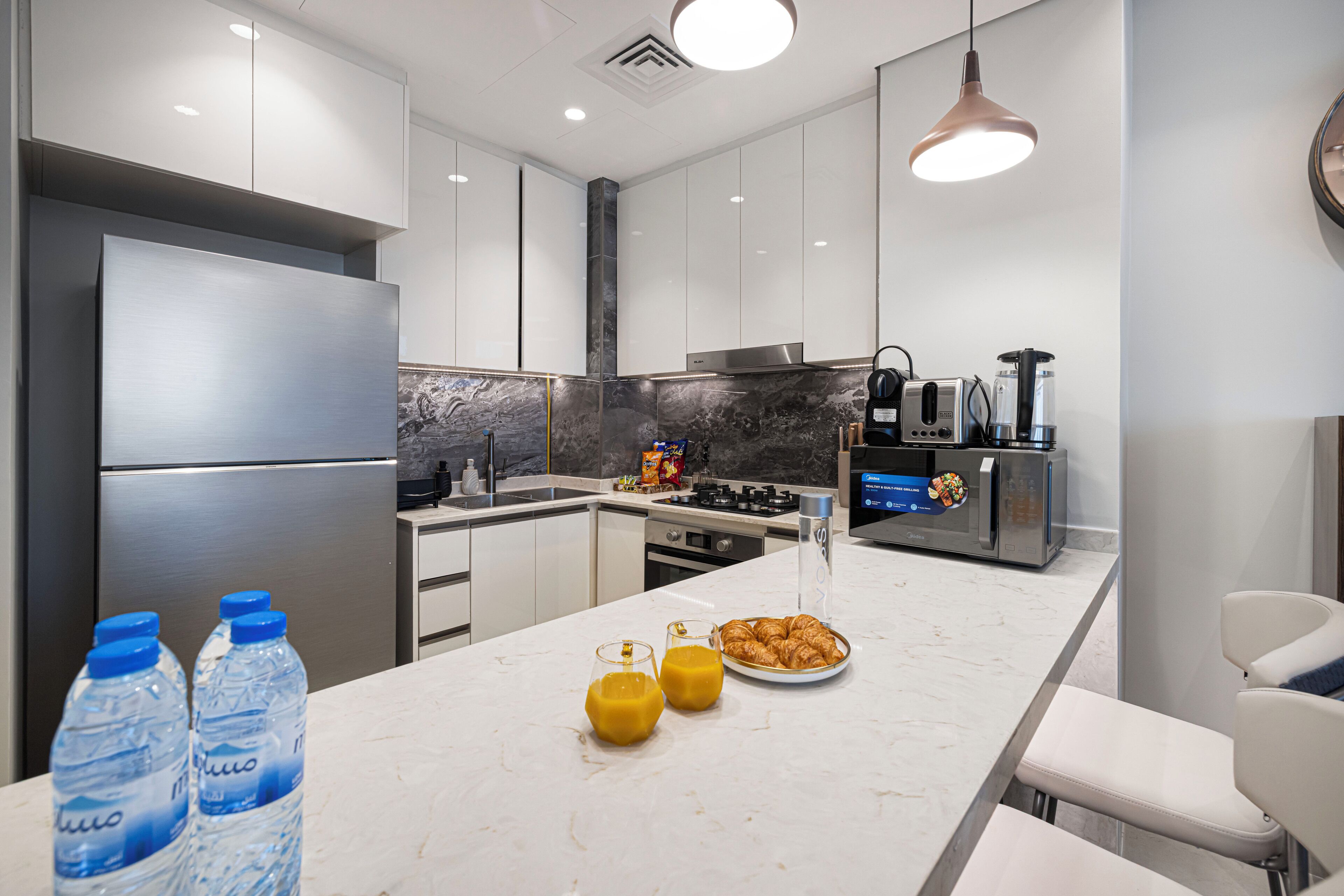 A modern kitchen with a stainless steel refrigerator, a marble countertop, and a breakfast setup including croissants and juice.